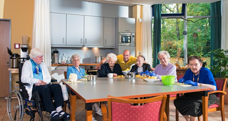 Bewohnerinnen und Bewohner mit Pfleger in der Küche des Altenpflegeheims sitzend und kochend